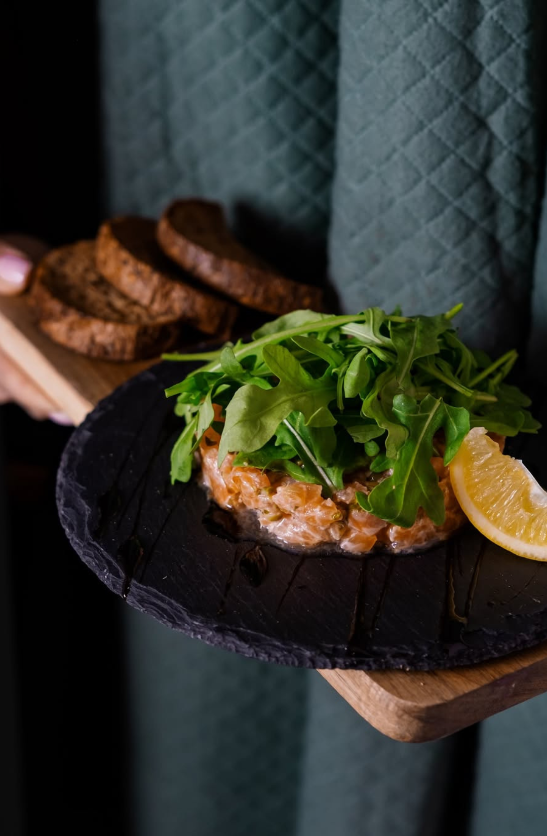 Salmon sandwich with arugula and a lemon wedge on a dark plate, held by a person.